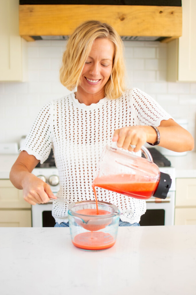 woman straining strawberry limeade through a fine mesh strainer at the kitchen counter.