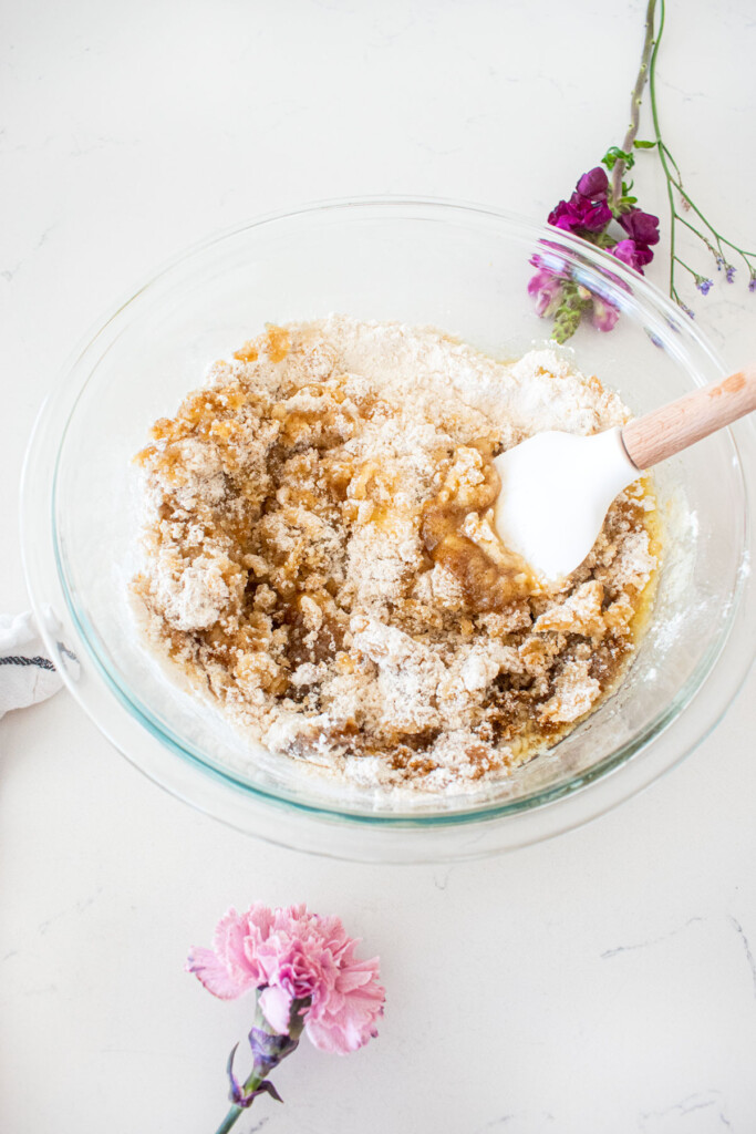 glass mixing bowl with ingredients being mixed with a spatula for cherry pie bars on a white marble counter with fresh flowers.