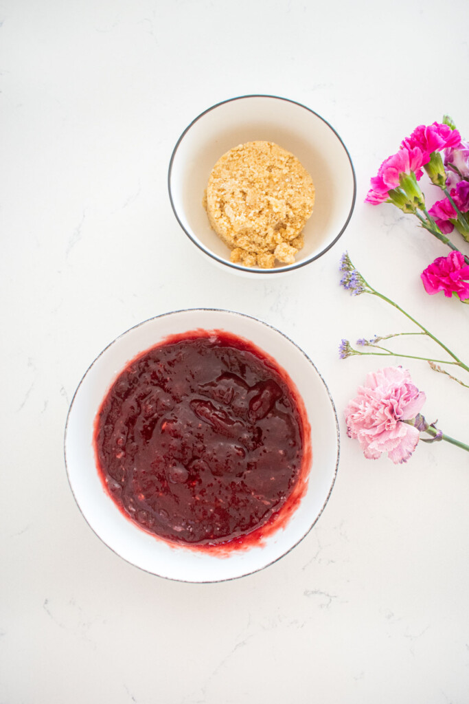 bowl of cherry jam and a bowl of dough in bowls on a white marble counter with fresh flowers.