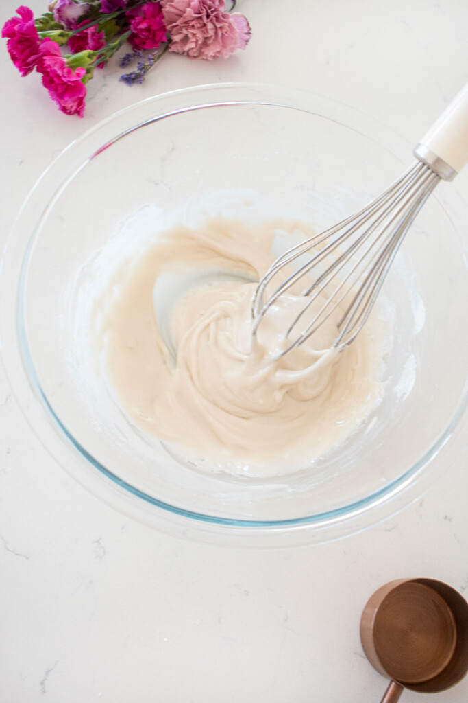 glass mixing bowl with glaze and a whisk on a white marble counter with fresh flowers.