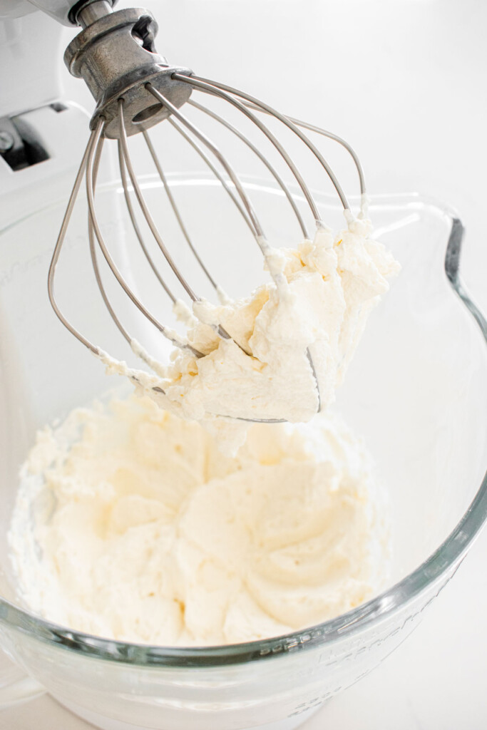 homemade whipped cream in a glass mixing bowl on a white marble counter.