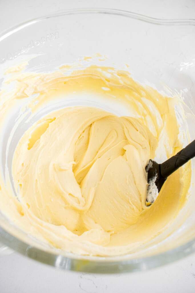 whipped cream being mixed into mango cheesecake filling in a glass mixing bowl on a white marble counter.