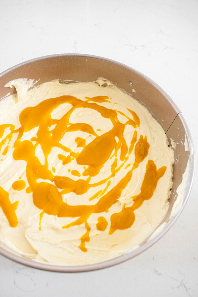 mango cheesecake with mango puree being swirled into it in a springform pan on a white marble counter.