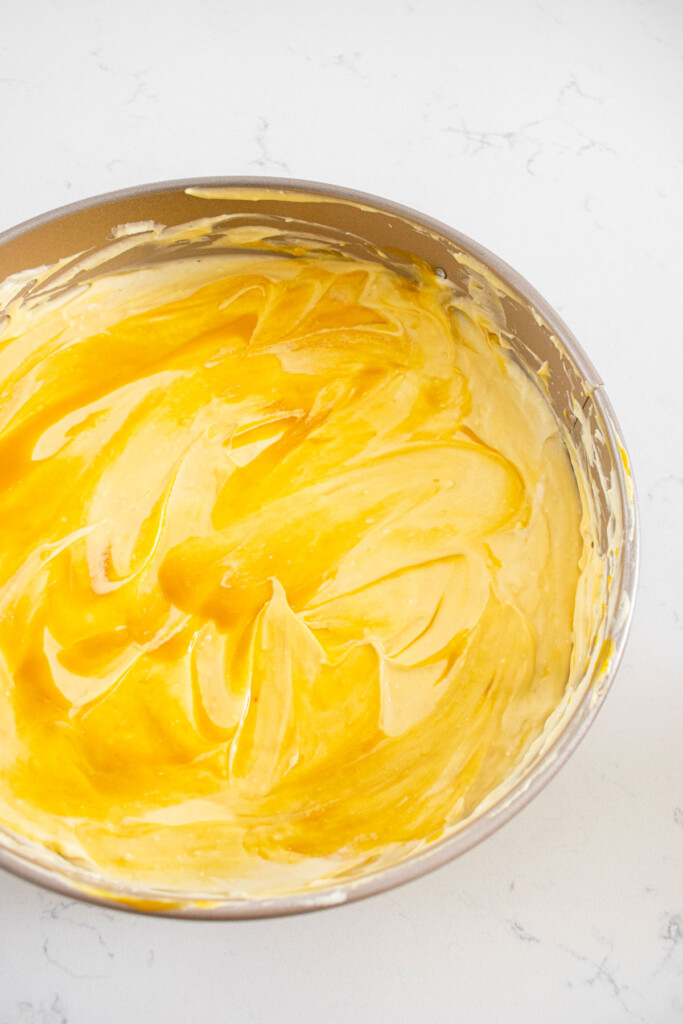 mango cheesecake with mango puree being swirled into it in a springform pan on a white marble counter.