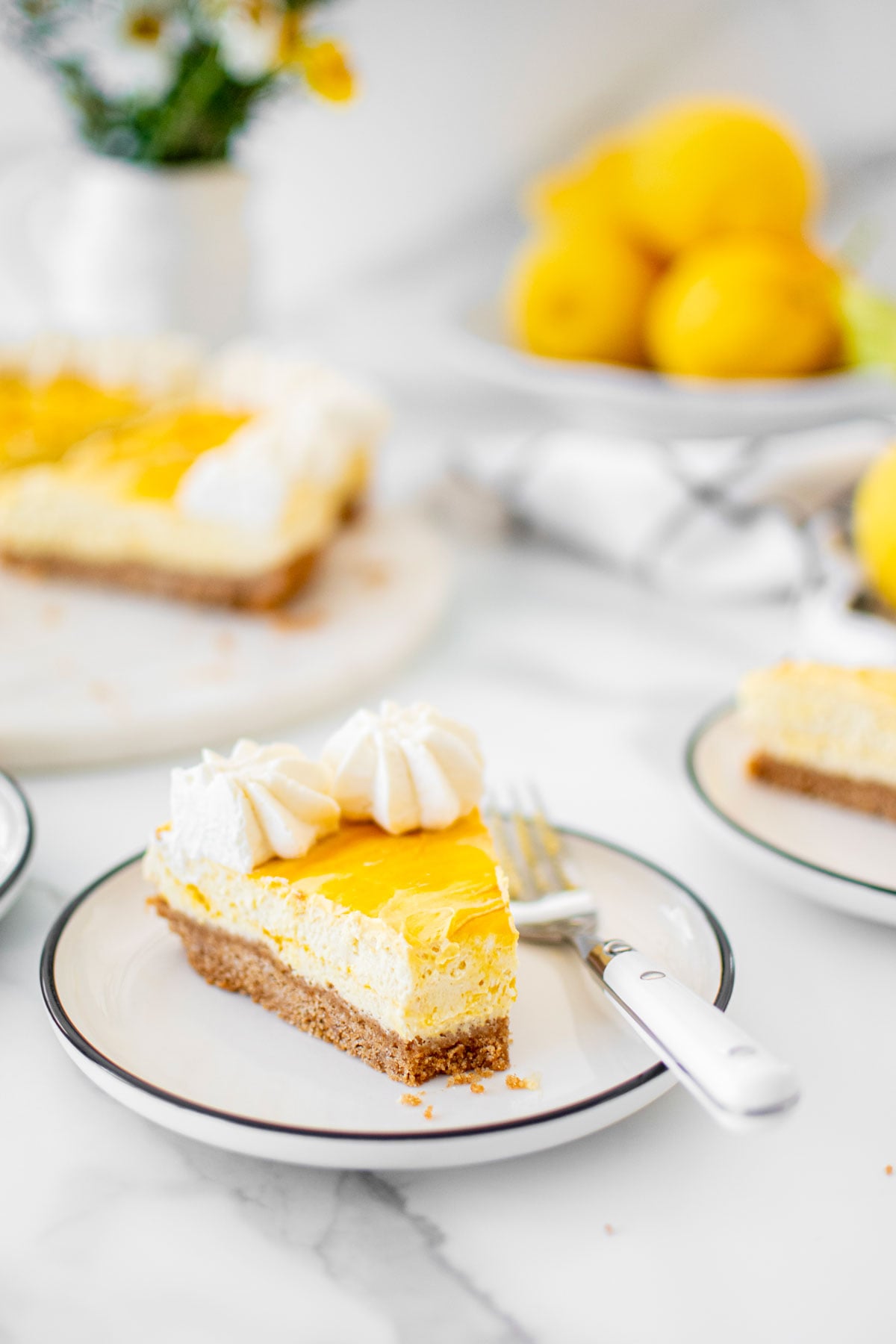 slice mango cheesecake on a plate on a white marble table with fresh lemons and flowers.