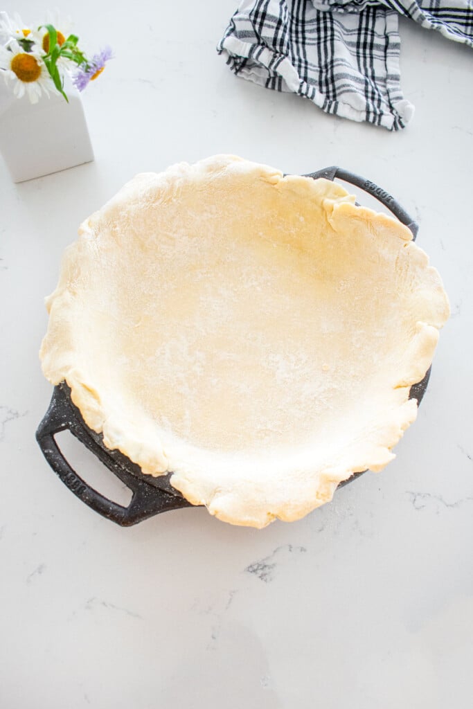 pie dough in a cast iron pie pan on a marble counter with a vase of fresh flowers.