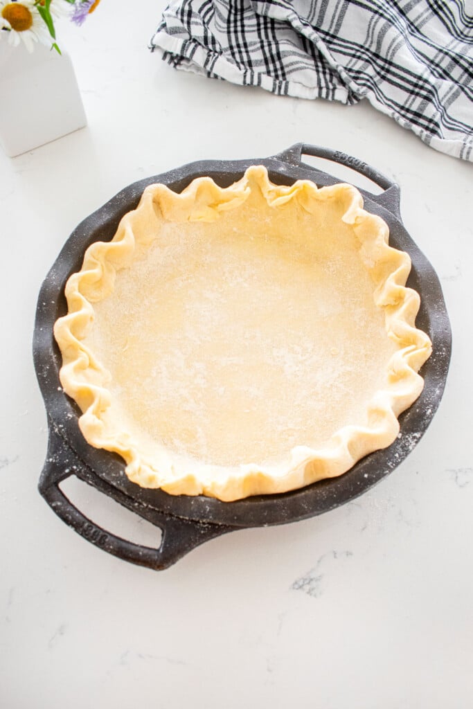 pie dough fluted in a cast iron pie pan on a marble counter with a vase of fresh flowers.