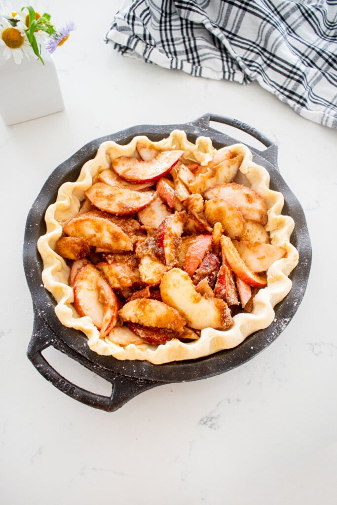 pie dough in a cast iron pie pan with peach pie filling on a marble counter with a vase of fresh flowers.