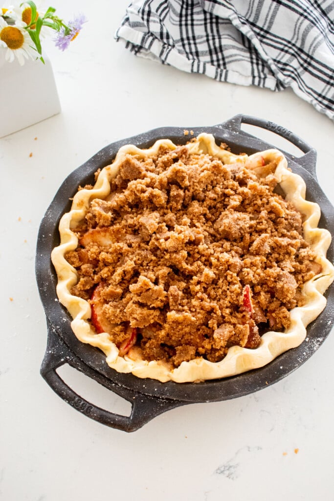 pie dough in a cast iron pie pan with peach pie filling and crumb topping on a marble counter with a vase of fresh flowers.
