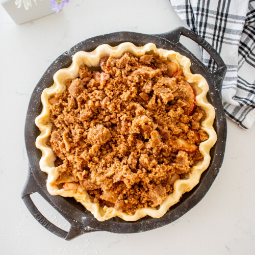 pie dough in a cast iron pie pan with peach pie filling and crumb topping on a marble counter with a vase of fresh flowers.