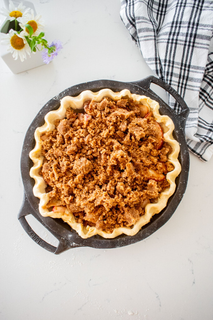 pie dough in a cast iron pie pan with peach pie filling and crumb topping on a marble counter with a vase of fresh flowers.