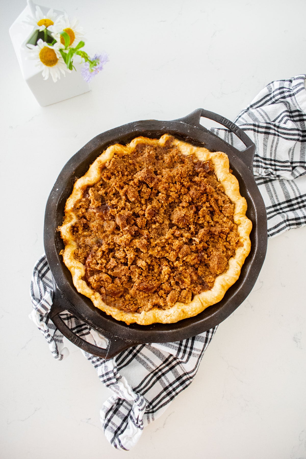 baked peach crumble pie on a marble counter on top of a plaid dish towel with a small vase of flowers.