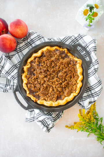peach crumble pie on top of a plaid dish towel on a table with fresh flowers and peaches.