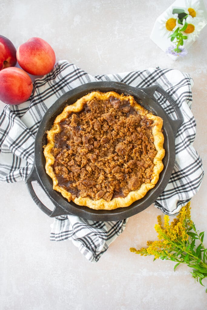 peach crumble pie on top of a plaid dish towel on a table with fresh flowers and peaches.