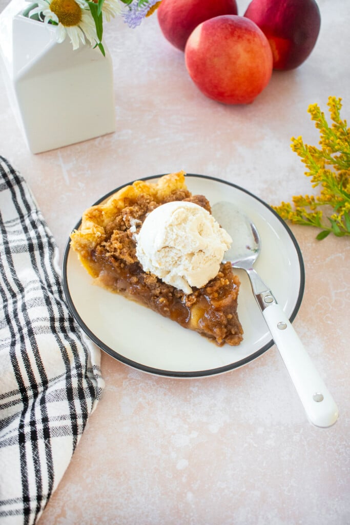 slice of peach crumble pie with vanilla ice cream on top on a plate on a table with fresh flowers.