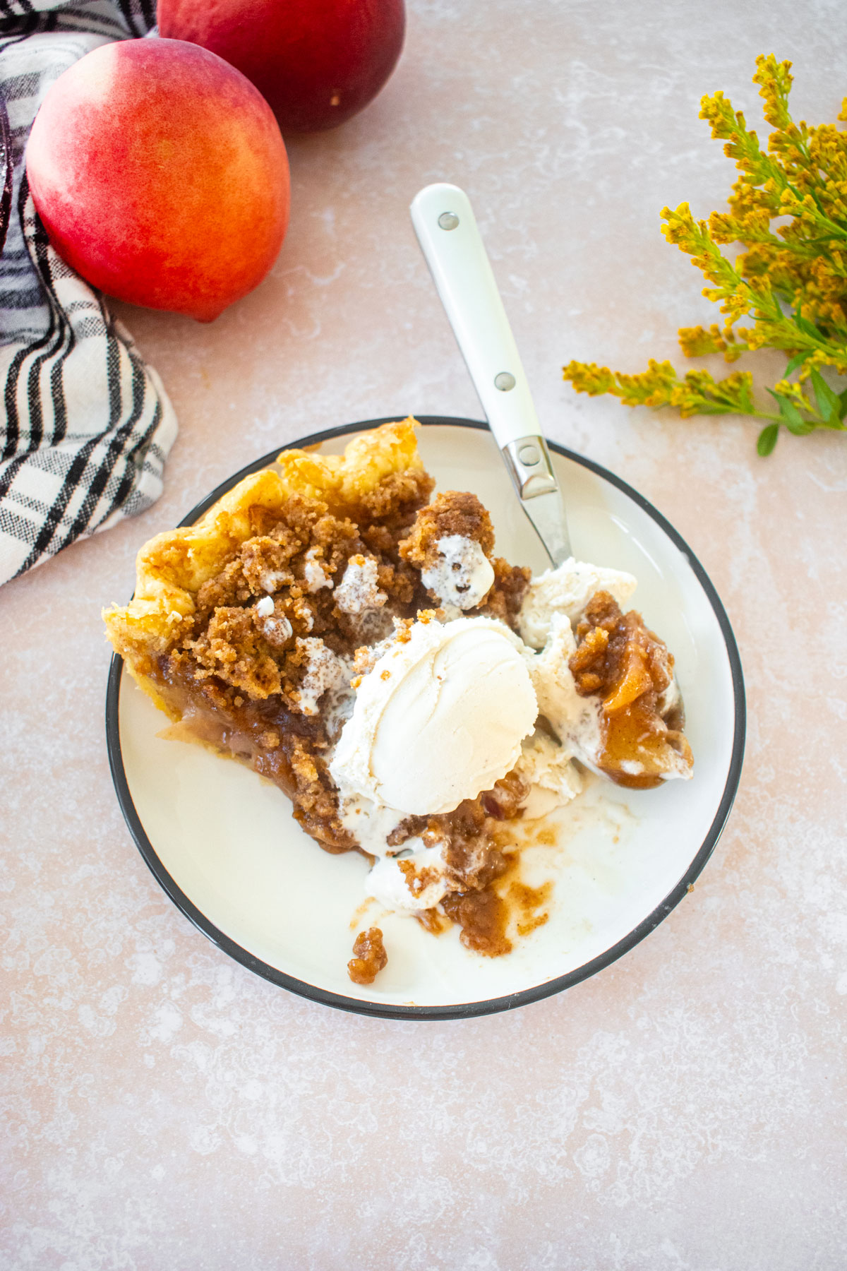 slice of peach crumble pie with vanilla ice cream on top on a plate on a table with fresh flowers.