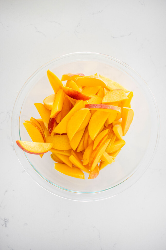 sliced peaches in a glass mixing bowl on a white marble counter.