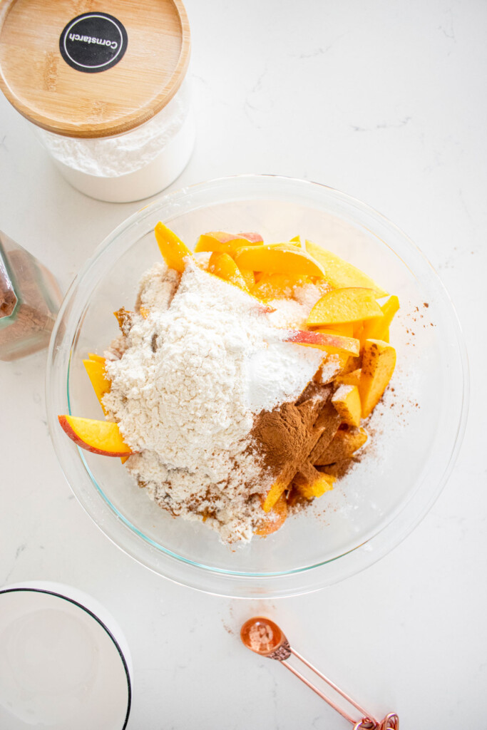 ingredients for peach crumble pie filling in a glass bowl on a white marble counter.