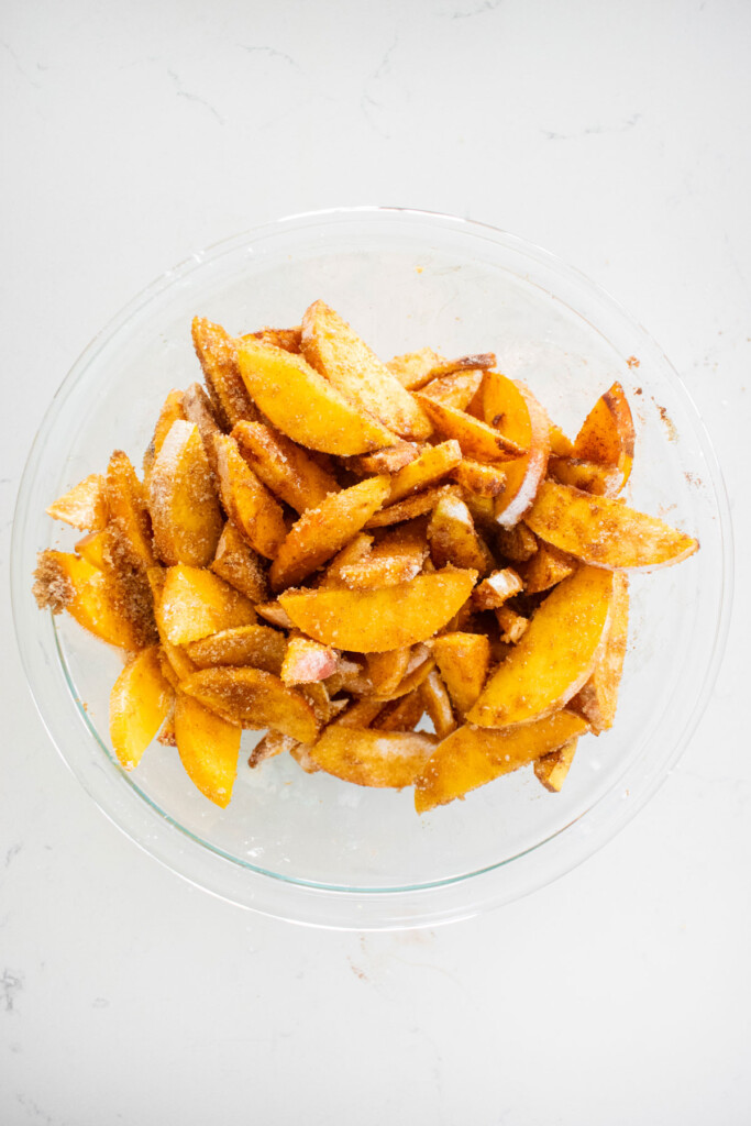ingredients for peach crumble pie filling in a glass bowl on a white marble counter.