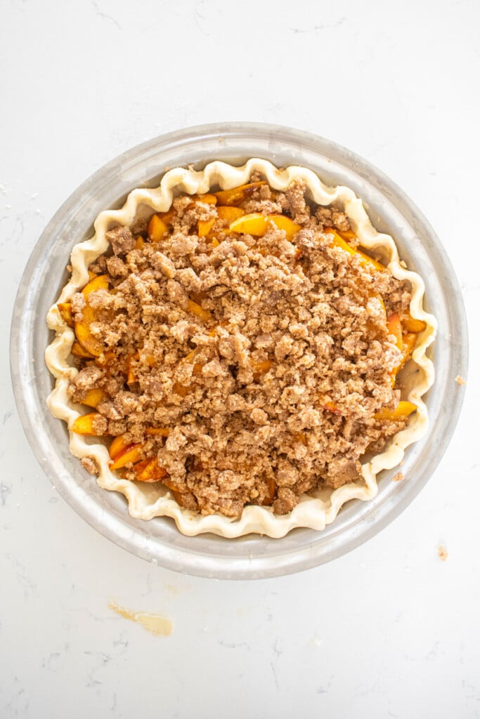 peach pie with streusel topping in a pie crust in a stainless steel pie pan on a white marble counter.