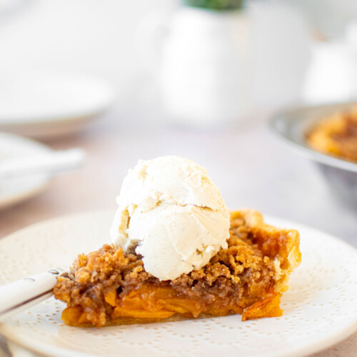 slice of peach crumble pie with a scoop of vanilla ice cream on a white plate.