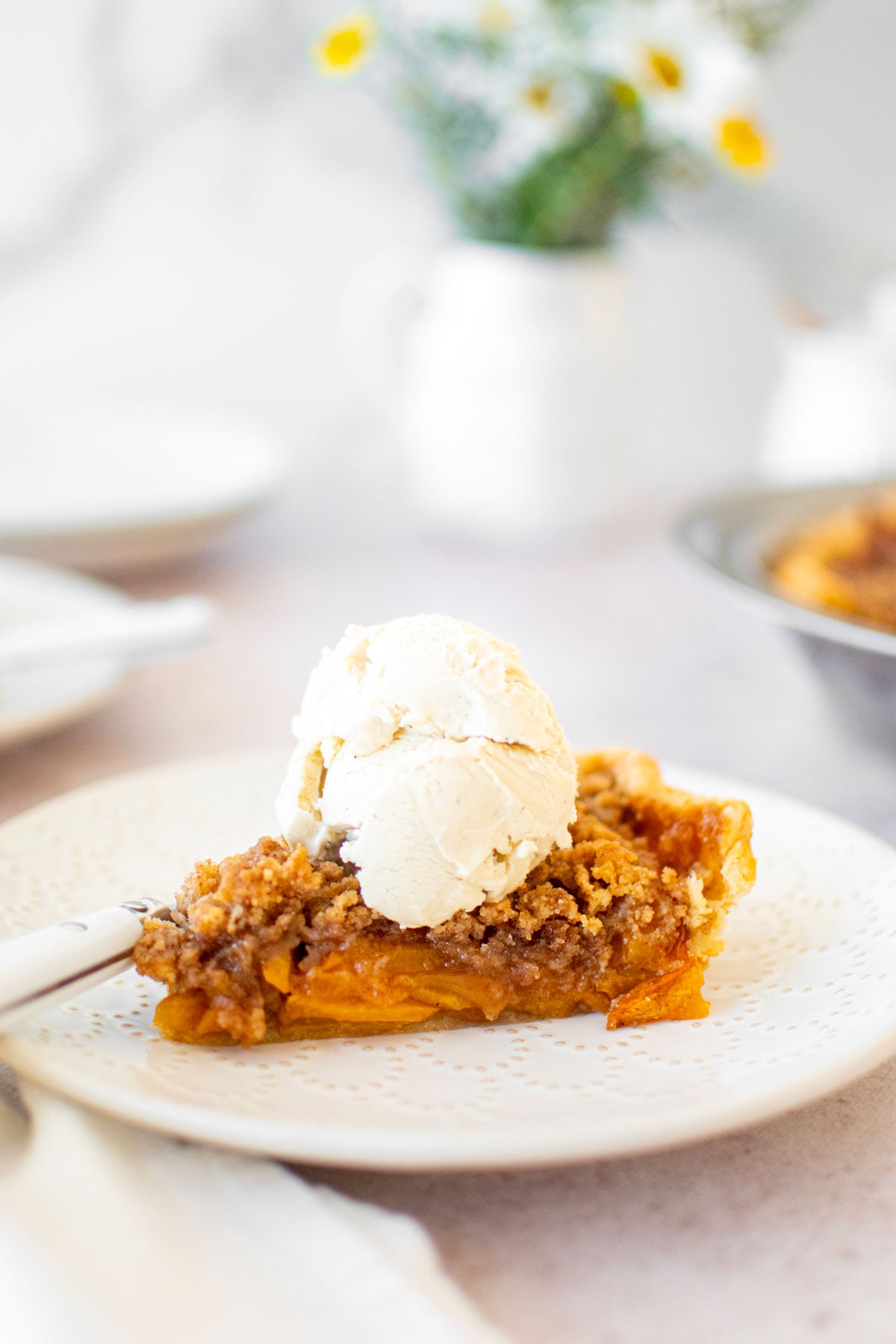 slice of peach crumble pie with a scoop of vanilla ice cream on a white plate.