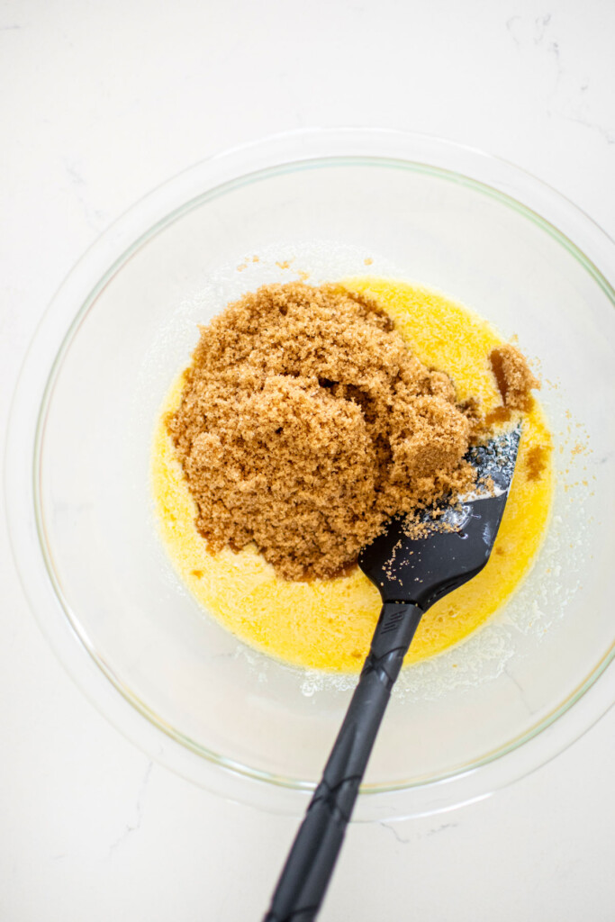 melted butter and brown sugar in a glass mixing bowl on a white marble counter with a spatula.