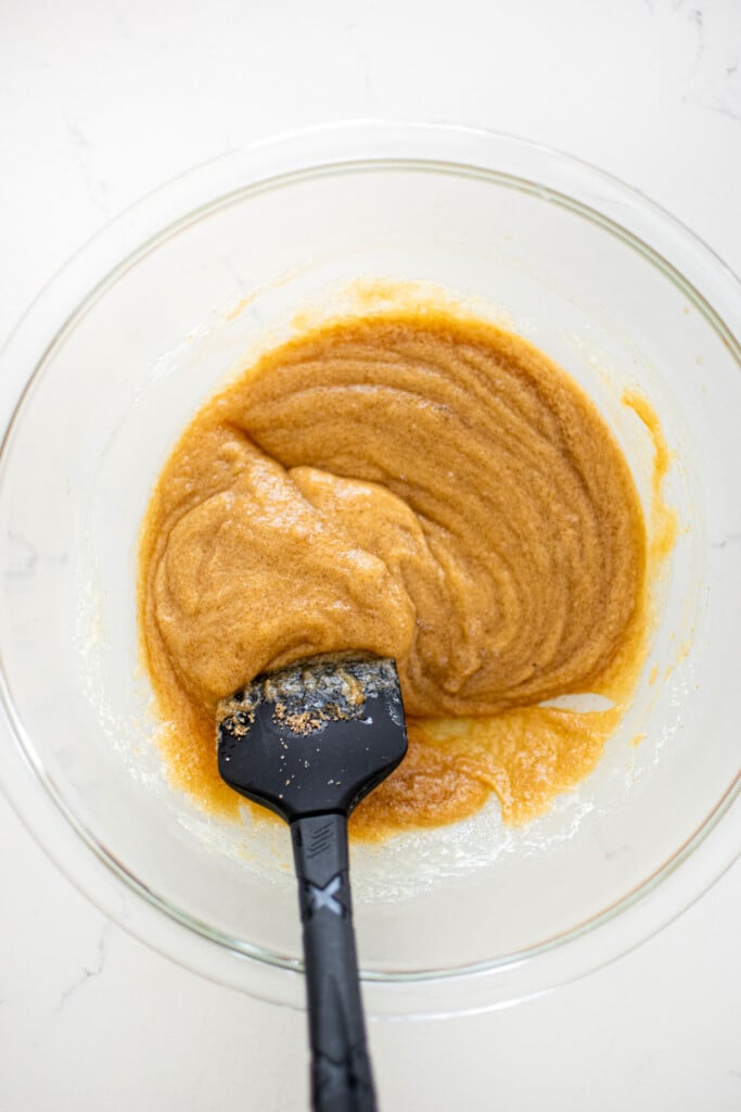 mixed melted butter and brown sugar in a glass mixing bowl on a white marble counter with a spatula.
