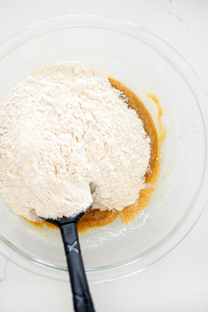 flour being added to cookie dough in a glass mixing bowl with a spatula on a white marble counter.