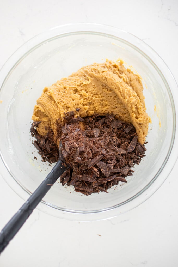 sourdough cookie dough with dark chocolate and a spatula in a glass mixing bowl on a white marble counter.
