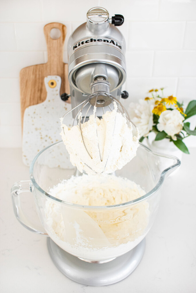 whipped cream whipped to stiff peaks in a glass stand mixing bowl on a white marble counter.