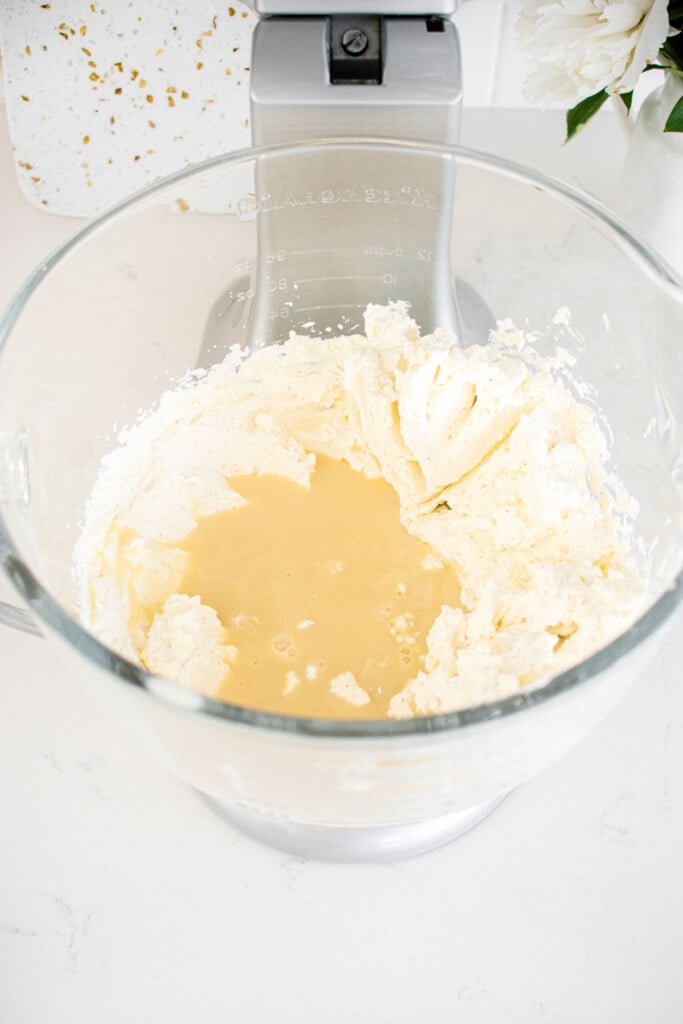 sweetened condensed milk added to whipped cream in a glass stand mixing bowl on a white marble counter.