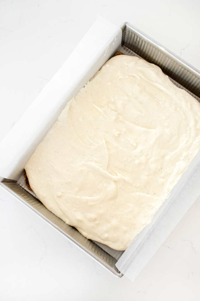 vanilla ice cream added into a baking pan on top of cookie layer on a white marble counter.