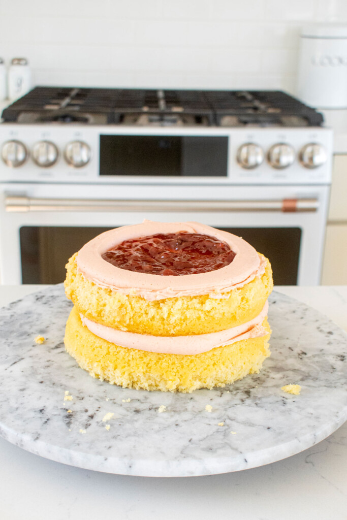 vanilla layer cake being assembled on a marble cake spinner with strawberry frosting and strawberry jam.