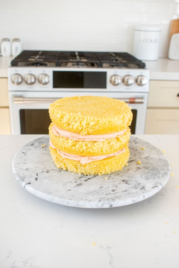 vanilla layer cake being assembled on a marble cake spinner with strawberry frosting.