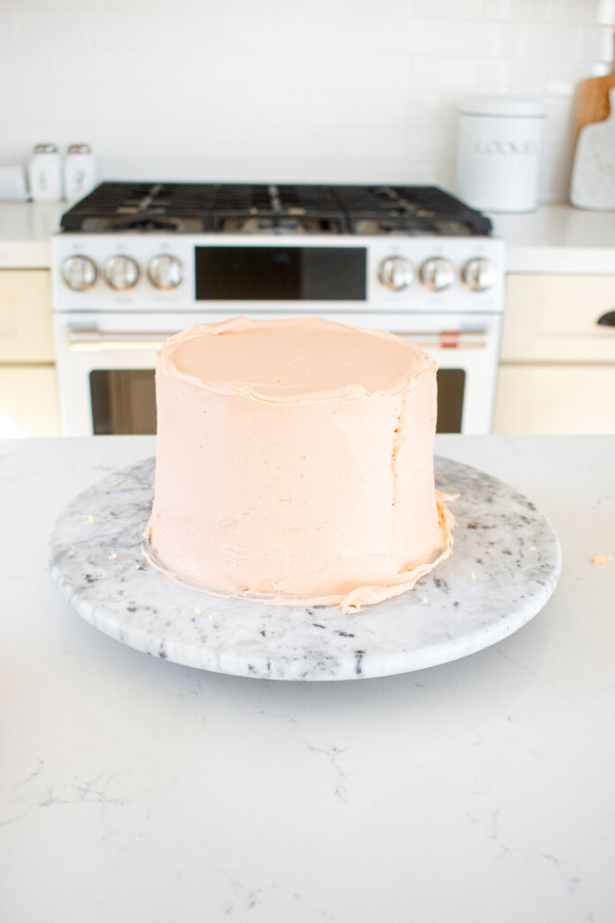 pink strawberry vanilla layer cake frosted on a marble cake spinner on the kitchen counter.