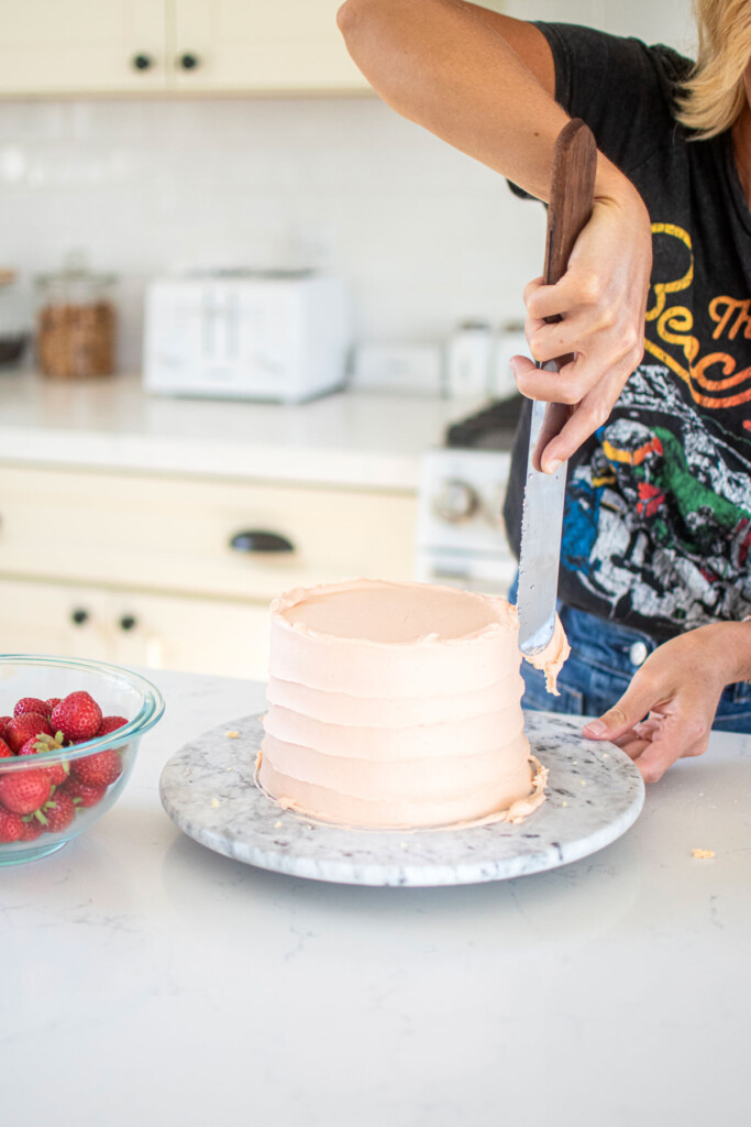woman frosting a pink strawberry layer cake on a marble cake spinner at the kitchen counter.