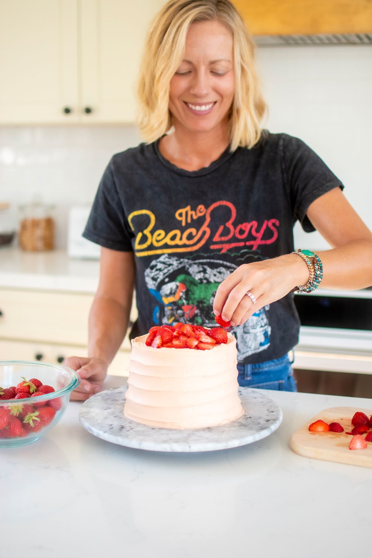 woman decorating a strawberry vanilla layer cake with fresh strawberries at the kitchen counter.
