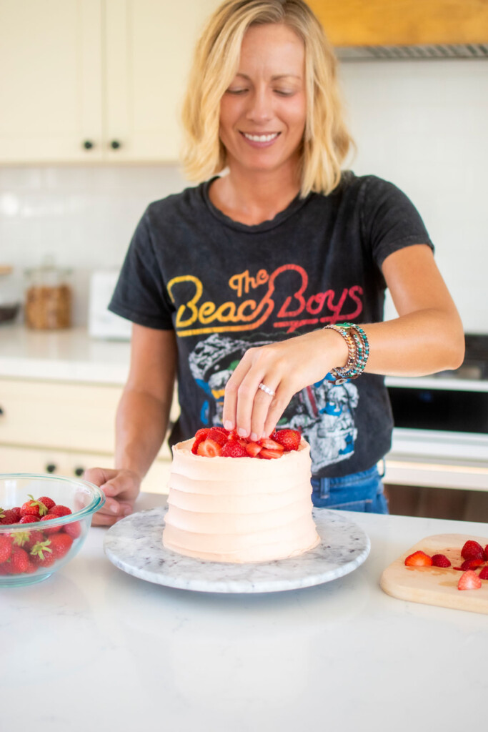 woman decorating a strawberry vanilla layer cake with fresh strawberries at the kitchen counter.