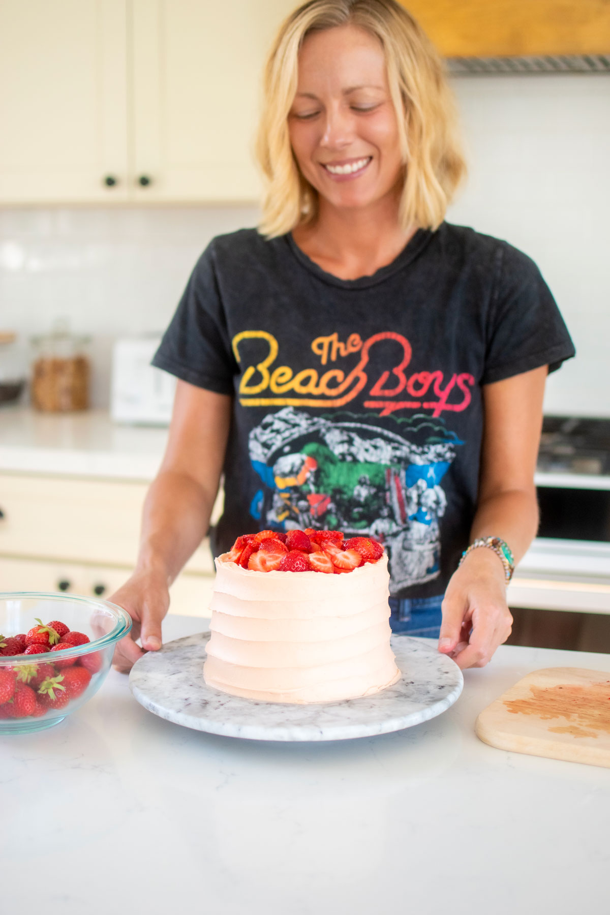 woman decorating a strawberry vanilla layer cake with fresh strawberries at the kitchen counter.