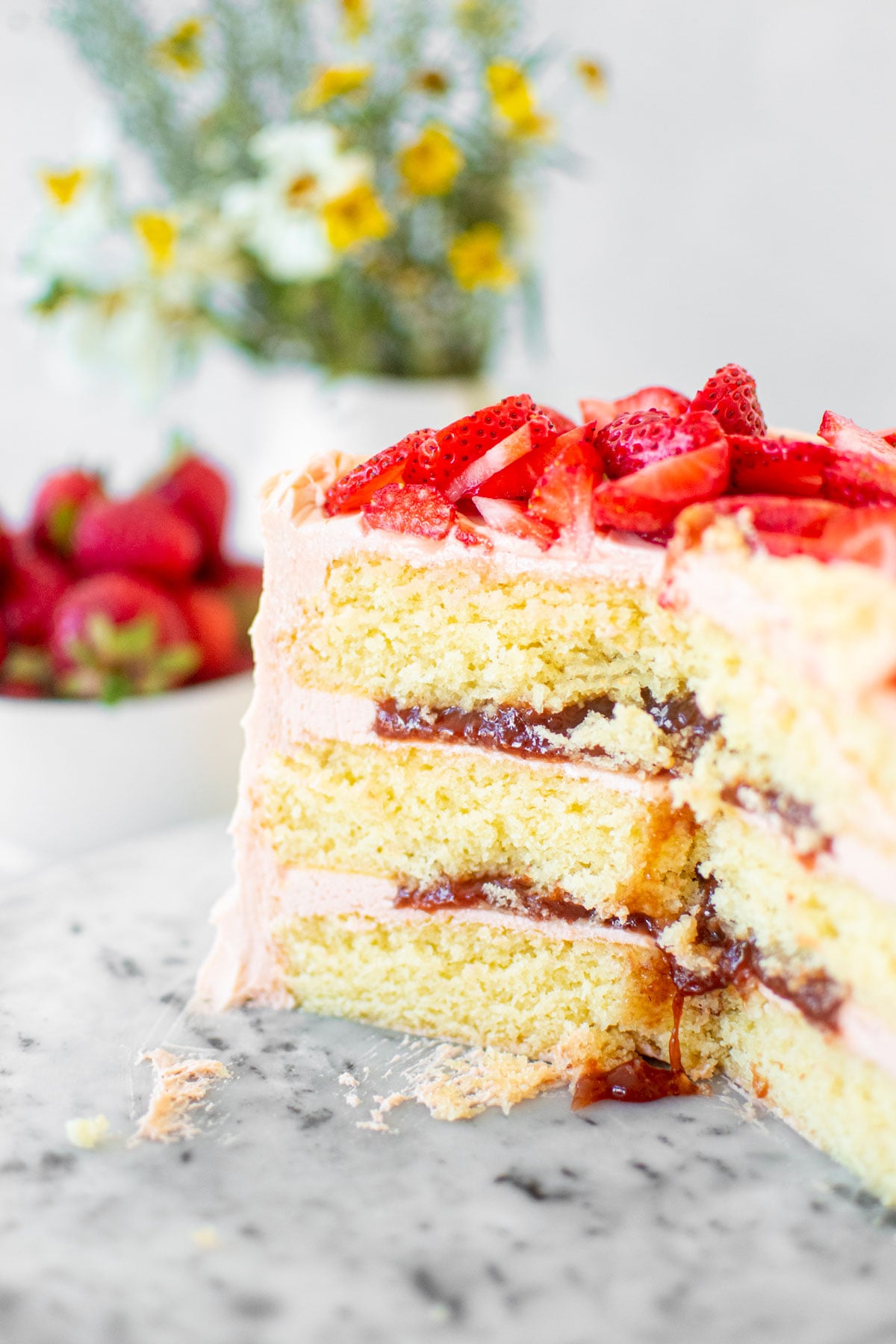 strawberry vanilla layer cake cut into on a marble cake plate to reveal strawberry jam filling with fresh strawberries and flowers in background.
