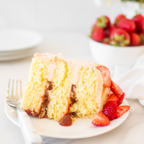 slice of strawberry vanilla layer cake on a white cake plate on a marble table with fresh flowers.