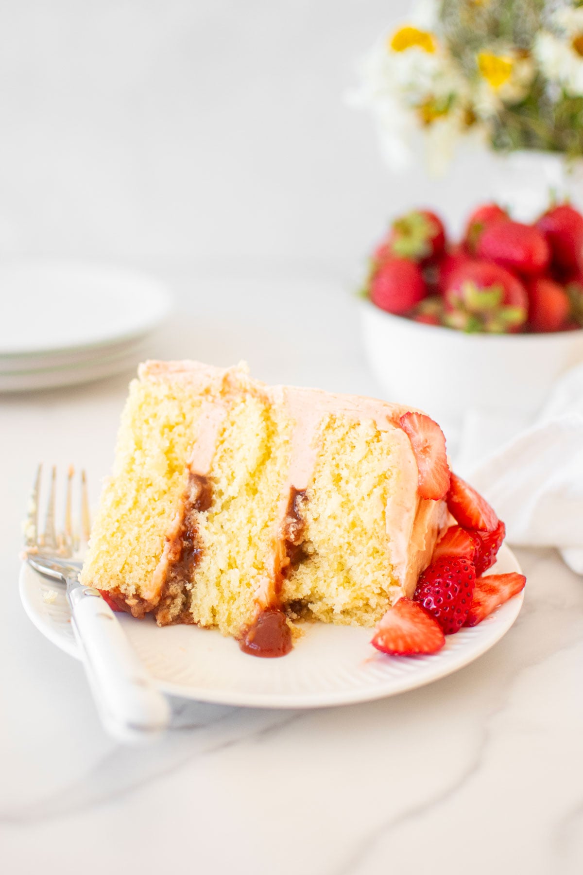 slice of strawberry vanilla layer cake on a white cake plate on a marble table with fresh flowers.