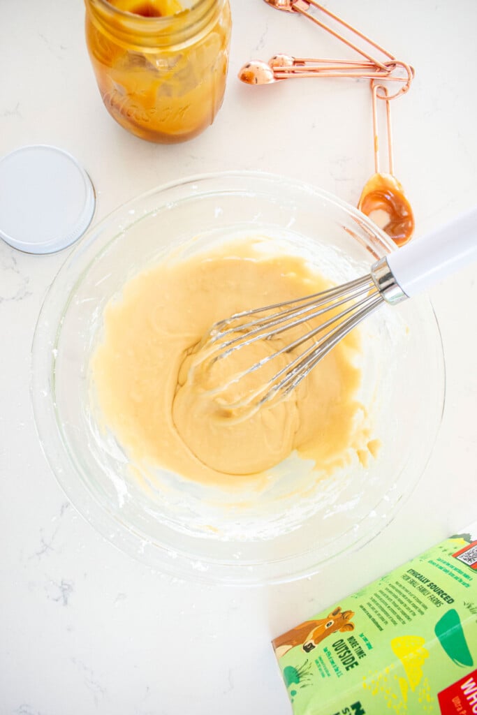 caramel icing mixed together with a whisk in a glass mixing bowl on a white marble counter.