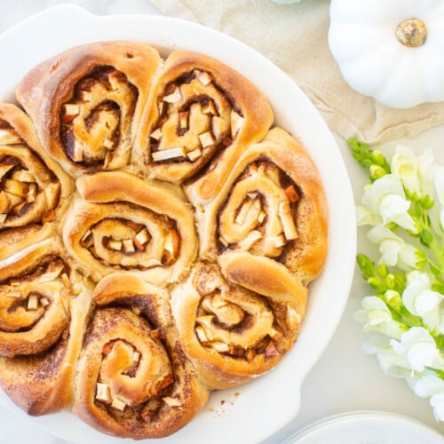 apple pie cinnamon rolls in a white pie dish on a white marble counter with pumpkins and fresh flowers.