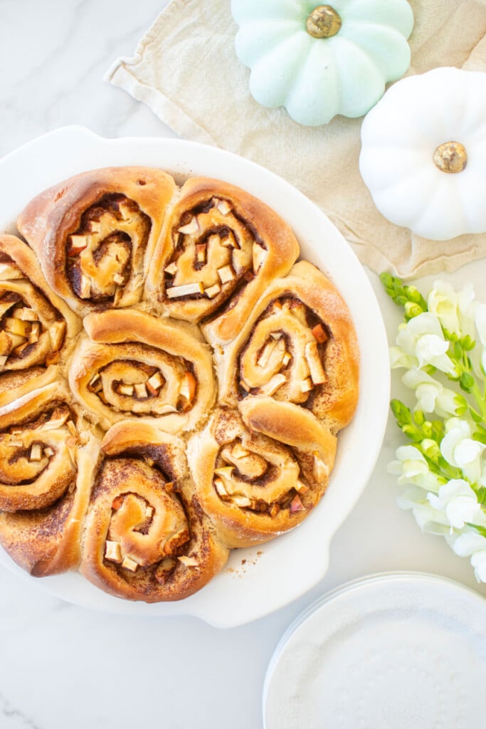 apple pie cinnamon rolls in a white pie dish on a white marble counter with pumpkins and fresh flowers.
