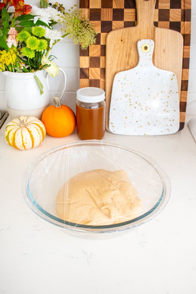 caramel apple pie cinnamon roll dough in a glass mixing bowl covered with plastic wrap on a marble counter with a jar of caramel and tiny pumpkins.