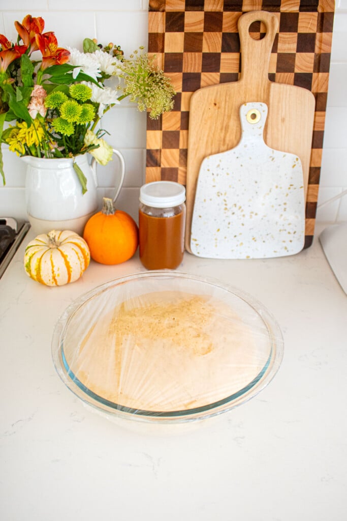 caramel apple pie cinnamon roll dough in a glass mixing bowl that has risen covered with plastic wrap on a marble counter with a jar of caramel and tiny pumpkins.