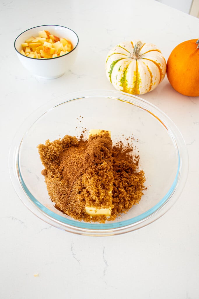 glass mixing bowl with butter, brown sugar, cinnamon and nutmeg on a marble counter with tiny pumpkins.