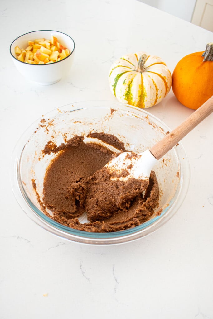 caramel apple pie cinnamon roll filling mixed together in a glass mixing bowl with a spatula on a marble counter with tiny pumpkins.